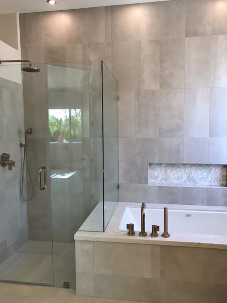 Image of a glass shower with tile on the walls. the entire back wall behind the shower and tub is tiled with gray rectangular tile. the tub is built-in and tiled around. there is a rain head for the shower head. Light is streaming in from a window on the right off the view.