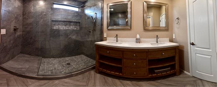 Panoramic of a remodeled upstairs master bathroom. Walk-in shower with gray 12x24 inch tiles, large wall glass panel, tiled niche, and window. His and Her sink vanity with center drawers and open shelves. over are personal framed mirrors. the floor is in a herringbone pattern with wood-like tile.