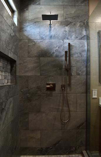 Image of a new tiled shower. The tile is a gray veined 12 by 24 inch tile. A long niche is on the left and a huge glass shower panel is on the right. Straight ahead is the rainfall shower head and the valve on the wall. there is also a handheld shower head with a slider. On the top left is a long short window that is streaming in the light.