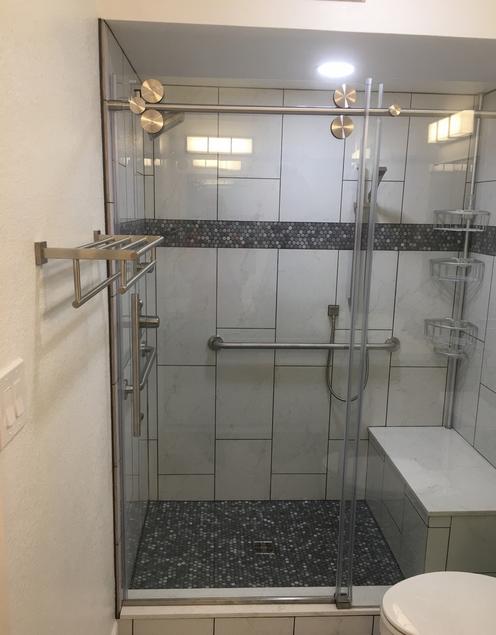 Image of a new tile shower with a glass barn-door style door. inside the shower is a tiled bench that matches the white rectangular tiles on the wall. there is a band of small glass mosaic tiles about 5 feet up the wall. the shower also has several mounted grab bars.