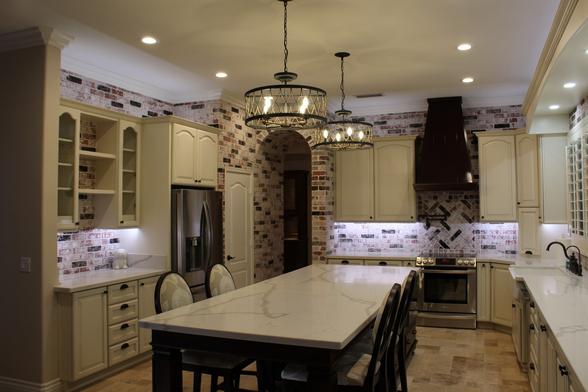 Image of a complete kitchen overhaul remodel. Faux brick on the walls and around the cabinets, with white grout that makes it look whitewashed. Huge kitchen island in the center with 4 seats around the end. White cabinets around the room, some with open shelves and glass doors with LED puck lights inside. A custom build walk-in pantry in the back corner of the room. The stove has a pot filler above it as well as a custom built copper finished large metal vent hood. The countertops are manmade white marble-looking.