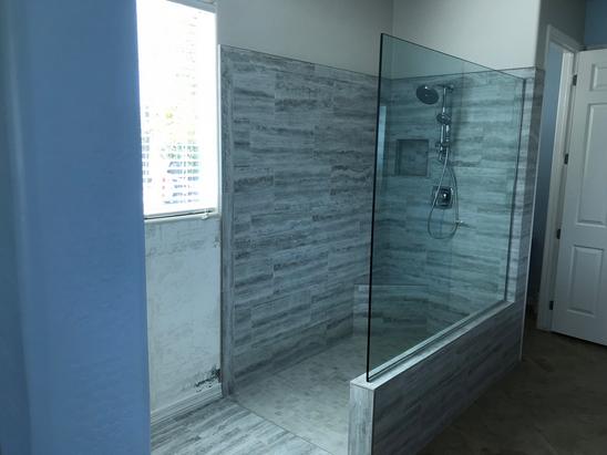 Image of a walk-in shower with a short pony wall with a large glass panel on top of it. The shower is tiled with gray and white rectangular tile and has a built-in corner bench as well as a small niche. light is coming in from a window next to the shower.