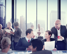 Corporate business meeting showing business executives in black business suits sitting and standing around boardroom table going over papers and spreadsheets.