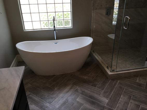 Master bathroom with gray herringbone floor tile. A large white oval freestanding tub and hand faucet. And a white marble tiled shower with glass panel and glass door
