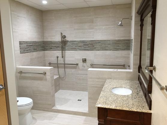 Image of a large walk-in shower with a pony wall on both the left and right of the shower entry. The tile is gray with some white veins and a glass tile band about 5 feet from the floor. There will soon be glass panels on both pony walls. In front of the shower to the right is a one sink vanity with a wood framed mirror. to the left is a toilet.