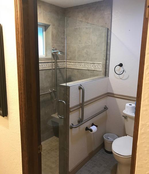 Image of an open doorway into a shower and toilet area. the shower has a pony wall with glass onto and a glass door to separate the toilet area. The tile in the shower is a tan 12 by 24 inch tile with a deco band of tile at chest height. There is a tiled floating bench on the shower wall and grab bars around the bathroom