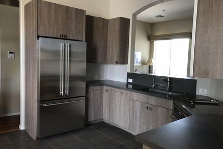 Picture of a newly remodeled kitchen. Gray cabinets with inset doors. Gray square tiles on the floor. The sink is right in front of a large wall opening that looks into the living room.