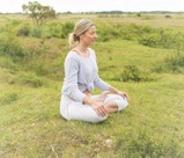 Blonde woman with pony tail sitting crossed legged in a grassy field meditating.