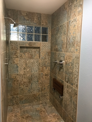 Walk-in shower with tiled linear drain and chrome fixtures. Unique gray, tan, and blue mosaic-like tile with a mounted teak foldable bench on the wall and a glass block window.