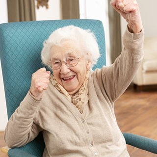 A smiling older woman sitting in a chair, raising her left hand.