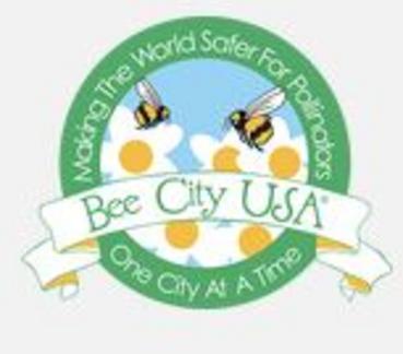 A circular Bee City USA logo with flowers and bees on it, saying "Making The World Safer For Pollinators, One City At A Time."
