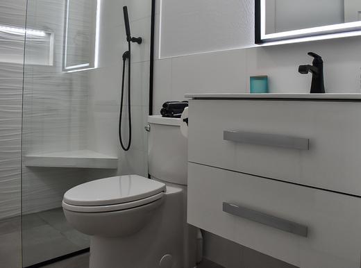 image of a newly remodeled second bathroom. in the foreground is a floating two drawer vanity with a black faucet and a framed LED mirror above. To the left of the vanity is a new toilet. To the left of that is a walk-in shower with a glass panel and a glass door. Inside the shower is white textured wavy 12 by 24 inch tiles. there is a bench in the corner under the shower head. On the wall is a niche with LED lights inside of it.