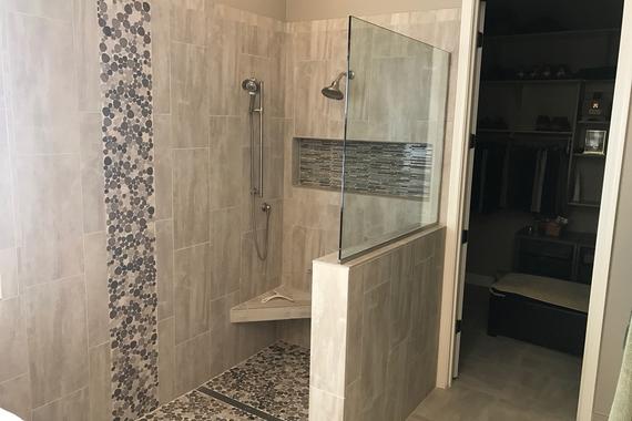 Master bathroom walk-in shower with a glass panel and mosaic river rock pebbles that resemble a waterfall going down the wall and onto the floor. Large niche, built-in mounted corner seat, and a linear drain.