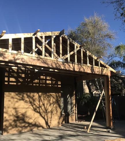 Image of a backyard patio with a built on patio cover being built. All of the framing, supports, and trusses are on.