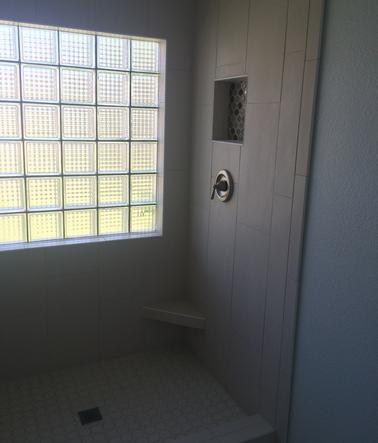 Picture of a shower without the glass shower door. On the back wall of the shower is a large glass block window. to the right of that is a floating corner shower bench. above that is the shower valve and niche.