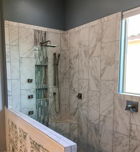 Image of a large walk-in shower with white tile with gray veins (looks like marble). There is a short pony wall that will soon have a glass panel on it. Under the shower head is a two tiered niche that has small glass tiles running from the top of the shower to the floor and looks like a waterfall. There are several body jets\sprays to the left of the niche. The light is coming in from a window on the right of the shower.