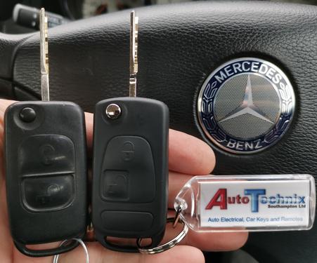 Picture of a Mercedes flip key and a replacement Mercedes key both black 2 button remote key in front of a Mercedes steering wheel with the Mercedes emblem in sliver and blue to the right of the picture