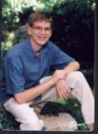 Senior picture of Andrew, outdoor with hand together with left forearm resting on his left knee, smiling, looking at camera.