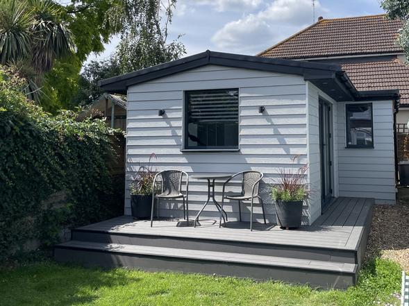 Grey slatted garden room with pitched roof and picture window with surrounding deck.