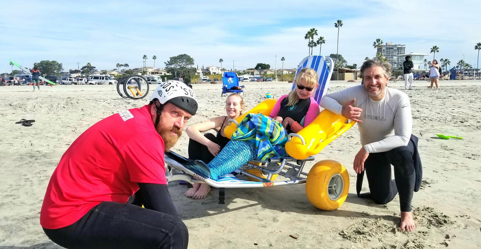 Picture of a people about to go surfing, a man holding a surfboard with a women holding his shoulder with a young girl all standing in the whitewater as waves roll in towards them.