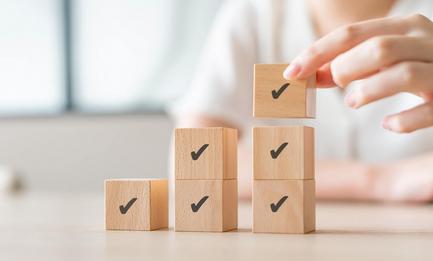 Three stacks of wooden blocks with checkmarks on a table and a hand placing one on top of the third column.