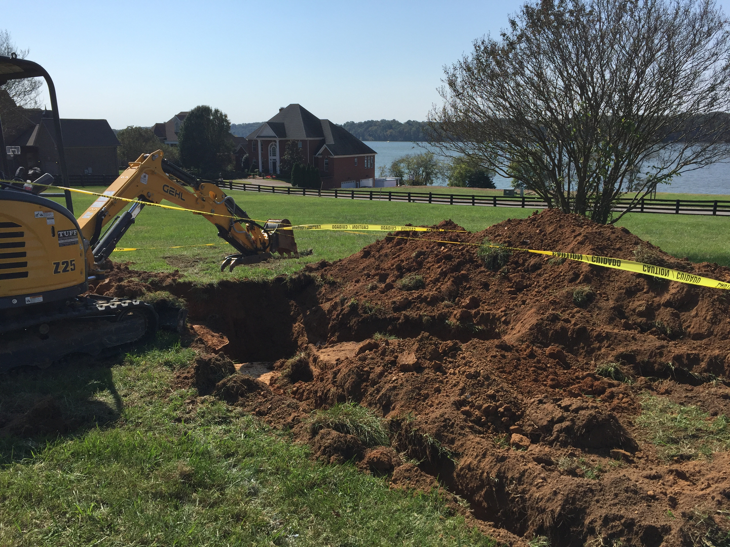 A construction site featuring an excavator digging into the ground, surrounded by mounds of soil. Caution tape is visible, marking off the area. In the background, there is a picturesque view of a lake and several houses.
