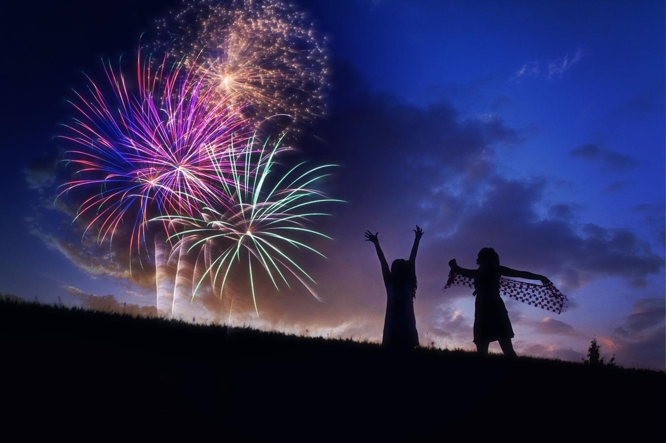purple, gold, red and green fireworks above silhouetted hillside with two silhouetted girls dancing and raising their arms