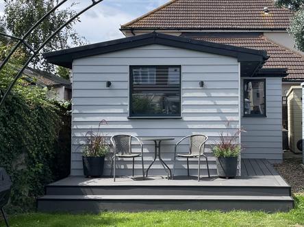 Grey slatted garden building with pitched roof and window with table and chairs on decking
