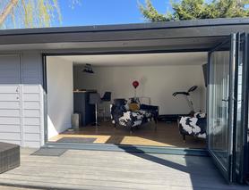 Grey clad garden room with open bifold doors showing a black and white sofa and chair and an exercise bike