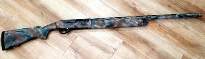Stoeger shotgun with "optifake timber" pattern lying on wood floor. Pattern is made of stencils with black, brown, gray, tan, beige and light grey.