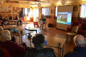 People sat in a semi circle behind tables, watching a presentation and speaker at the centre.