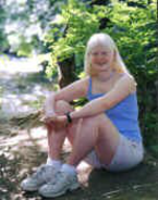 Outdoor picture of Caitlin, sitting with back against a rock, arms on knees, smiling looking at camera.
