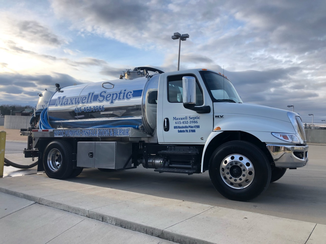 A large septic service truck with a shiny silver tank and blue hoses, parked on a concrete surface under a cloudy sky. The truck features the name Maxwell Septic prominently displayed on its side, along with a contact number and a description of septic pumping, installation, and repair services.