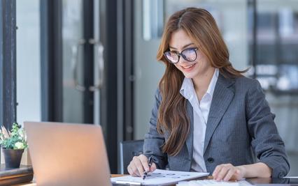 Image of a young lady working at an office desk, with reports, and laptop