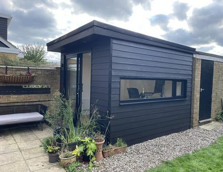 Small garden office clad in black weatherboard with sliding doors at the front and a long horizontal window
