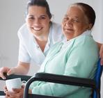 Image of older female adult sitting in wheel chair smiling, Women is accompanied by female medical staff also smiling.
