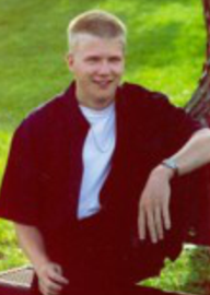 Senior picture of Kale, sitting, left elbow on tree, smiling at camera.