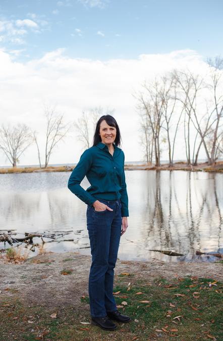 Alison L Randall stands in front of a pond and a lake is visible in the background. She wears a teal blue shirt and jeans