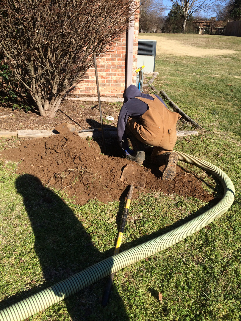 A person wearing a hooded sweatshirt and overalls is crouched down digging in the soil near a brick wall. A shovel and a green hose are visible in the foreground, with a bush and grass surrounding the scene. The background shows a grassy area with trees and a building.