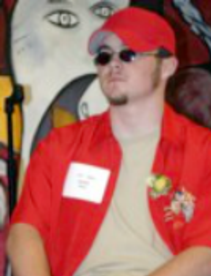 Kenny, sitting on chair at 2004 STF reception, wearing sunglasses and a bright red hat and shirt, looking to his right.