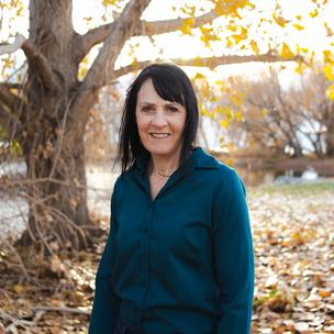 Alison L Randall wears a teal blue shirt and stands in front of a golden leaf-covered patch of grass. A pond peeks out in the distance