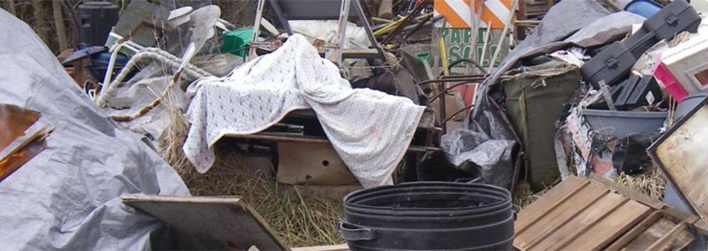 A background banner photo of a yard full of junk sitting in heaps consisting of plastic cases, tarps, and wood pallets, along with much, much more to clutter up the image. A hint of fire is just off to the left of the photo-- A hint of transformation in a world overcome with garbage. (Photo was taken by the owner years before starting the business as part of his ongoing interest in positive transformation through smart, well done, and creative work.)