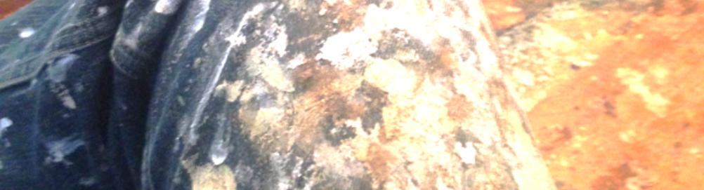 A photo banner background of a closeup of Nathan's painting jeans-- A dark blue denim covered in many energetic splotches of white and orange paints. The background is also very textured, an image of a painter's floor, covered in yellow and orange splotches. The whole image is in slightly elevated contrast and a bit faded, like a slightly distorted newspaper photo.