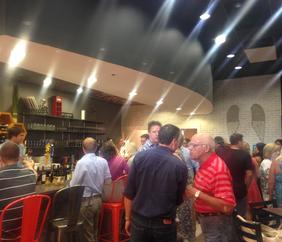 A bustling grand opening of a brand new wine bar and boutique, recently remodeled in time for the big day. A dark grey painted overhang with can lights casts streaks of light across the photo as a group sits in red and black chairs admiring the wine selection and several other groups gather to talk around the boutique area, a greenish-white faux brick wall (Completed by 180 Improvements) with a set of matching decorative wings makes up the background.