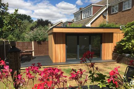 Modern cedar clad garden room with 3 panel bifold doors viewed through red flowers
