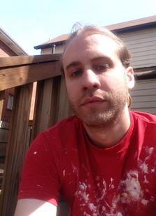 A photo of the owner, Nathan, from the chest up, sitting on the steps of a deck in a backyard in St. Louis city. He has blondish brown hair, blue-green eyes, a very short beard, hair in a ponytail, and is wearing a red shirt with splotches of white paint on it, deck railing just visible over his right shoulder and a house with tan siding, a brown roof, and dark brown gutters in the background set under a bright, light blue sky.