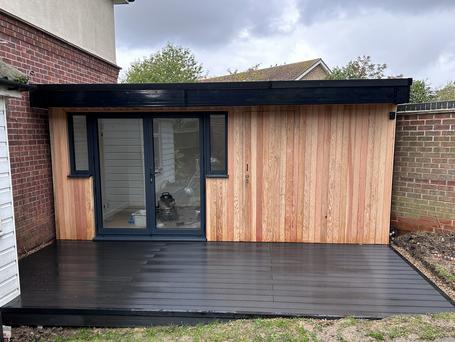 Larch clad garden building with dark grey French doors and small windows either side with a large black decking area in front