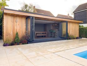 Cedar clad garden room in front of an outdoor pool with open bifold doors showing a bar, green bar stools and a table