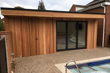 Modern cedar clad garden room pool house with 3 panel bifold doors and integral shed next to an outdoor swimming pool