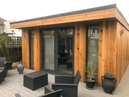 Modern cedar clad garden room with French doors and 2 full length windows either side on a decking area with tables and chairs